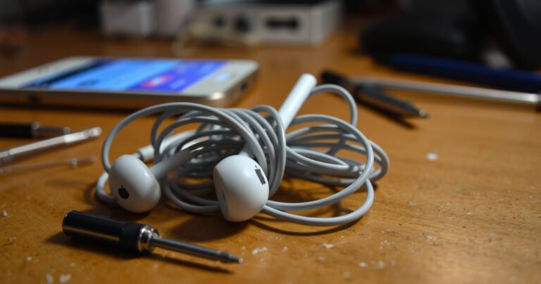 A close-up view of a pair of tangled Apple earphones on a wooden table, surrounded by scattered tools like a tiny screwdriver and a pair of tweezers, with a faint glow of a smartphone displaying troubleshooting tips in the background, evoking a sense of DIY repair and technology in action.
