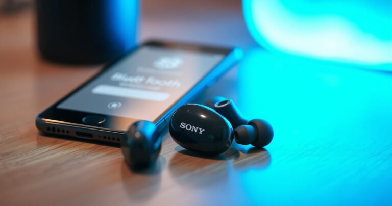 A close-up of sleek Sony earbuds resting on a wooden surface, surrounded by soft, glowing blue light. Nearby, a smartphone displays a Bluetooth pairing screen, with soft reflections highlighting the earbuds' glossy finish. The background is blurred, evoking a sense of modern technology and effortless connectivity.