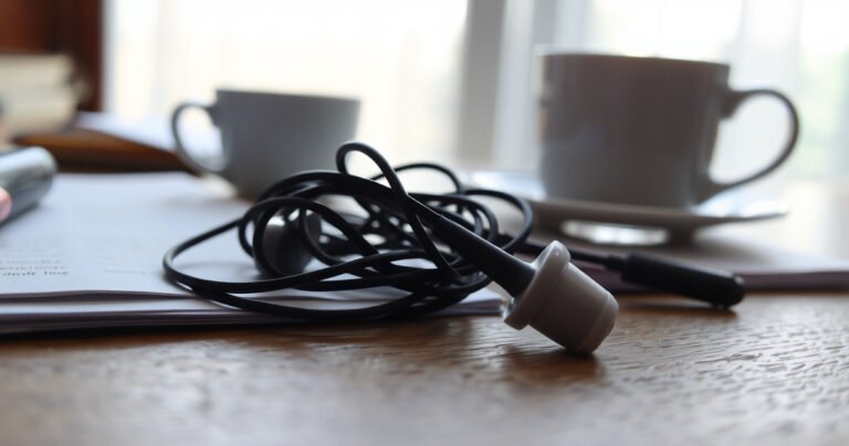 A close-up of a tangled pair of earbuds resting on a wooden table, one earbud glowing faintly while the other lies dull and silent. Surrounding them, scattered notes and a half-empty coffee cup, with soft morning light streaming through a nearby window, creating an atmosphere of curiosity and frustration.